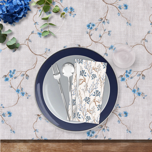 Navy blue plate with silverware and floral napkin on a decorative tablecloth with blue flowers and green leaves.