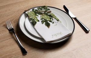 Table setting with a white plate, green leafy napkin, and silverware on a wooden surface.