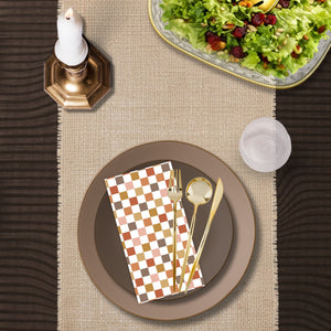 Top-down view of a taupe and brown disposable table setting with gold plastic cutlery, patterned paper napkin, plastic plate, woven placemat, salad bowl, and candle centerpiece.