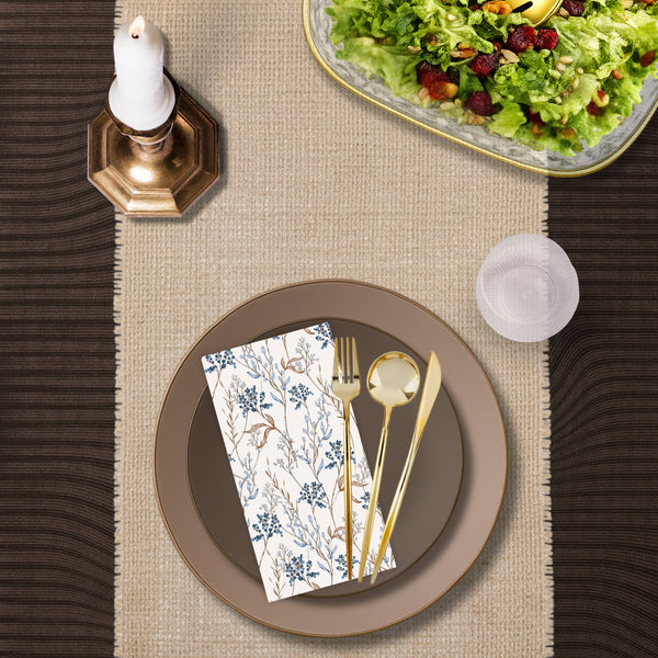 Top-down lifestyle image of a taupe and brown disposable dinnerware place setting with gold plastic cutlery, blue floral paper napkin, clear plastic goblet, woven runner, candle, and salad bowl.