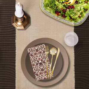 Top-down view of a taupe and brown disposable dinnerware place setting with layered plastic plates, brown floral paper napkin, gold plastic flatware, and a clear plastic goblet on a woven table runner.