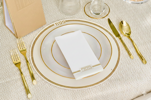 Elegant table setting with gold-rimmed plates, gold cutlery, and a white napkin on a textured beige surface.
