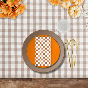 Fall-themed disposable dinnerware place setting with orange plate, taupe charger, gold flatware, ribbed goblet, and checker print napkin on a beige gingham tablecloth.