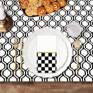Top view tablescape featuring white and gold scalloped plastic plates, gold flatware, clear goblets, and black and white checkered napkins on a geometric tablecloth