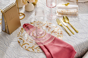 Elegant table setting with gold cutlery, pink napkin, and decorative glassware on a white lace tablecloth.