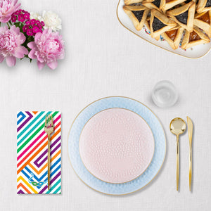 a purim tablescape with Colorful striped purim themed napkin, pink and blue plates with gold rim, gold cutlery, and floral arrangement on a white table.