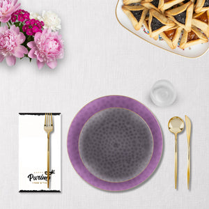 Table setting with purple and gray plates, gold cutlery, and flowers on a white tablecloth.