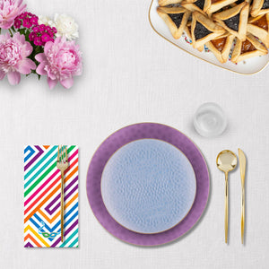 Colorful purim table setting with a purple plate, gold cutlery, and a rainbow striped napkin.